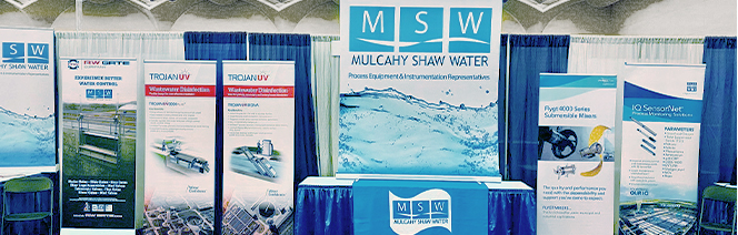 A trade show booth for Mulcahy Shaw Water features banners, equipment images, and informational displays about water treatment solutions, with a central blue table and backdrop displaying the companys name and logo.