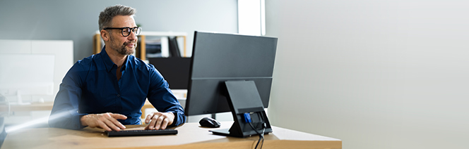 A man with glasses and a blue shirt sits at a desk, working on a large computer monitor in a modern, bright office space.