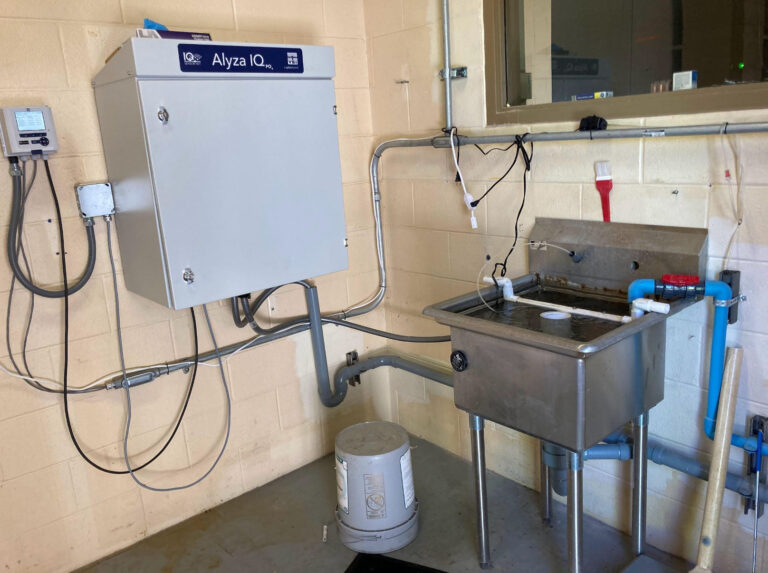 A laboratory sink area with a stainless steel sink, pipes, a control panel labeled Alyza IQ, cleaning tools, and a large white bucket on the floor, set against a beige tiled wall.