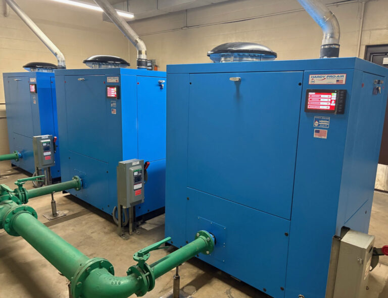 Three large blue industrial machines with control panels and ventilation pipes are lined up in a utility room, with green pipes connected at the front and a beige wall in the background.