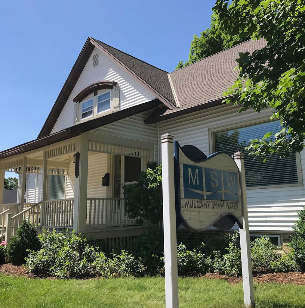 A white house with a covered porch, surrounded by green shrubs and grass. In front, there is a blue and white sign that reads “MSW Mulcahy Shaw Water.” The sky is clear and sunny.