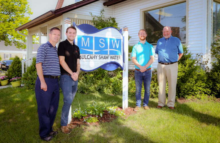Four men stand smiling in front of a Mulcahy Shaw Water (MSW) sign outside a white building on a sunny day, with greenery and flowers around the sign.