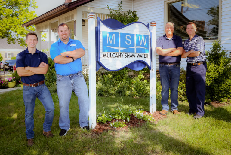 Four men stand smiling with arms crossed around a sign that reads MSW Mulcahy Shaw Water in front of a white building and green landscaping on a sunny day.
